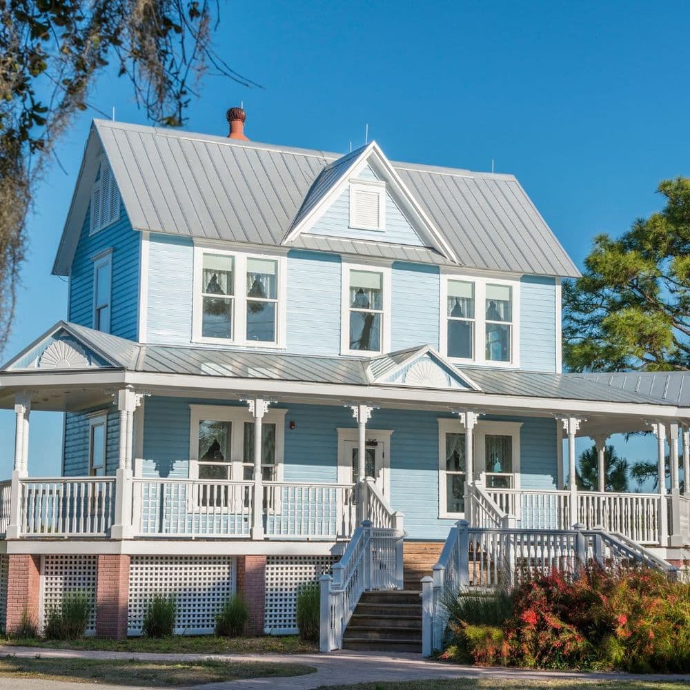 Blue Victorian-style house with white trim and a spacious front porch surrounded by greenery.