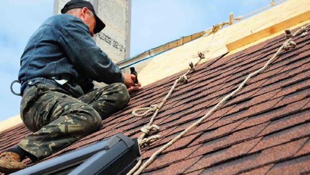 Roofer installing shingles on a residential roof, secured with safety ropes.